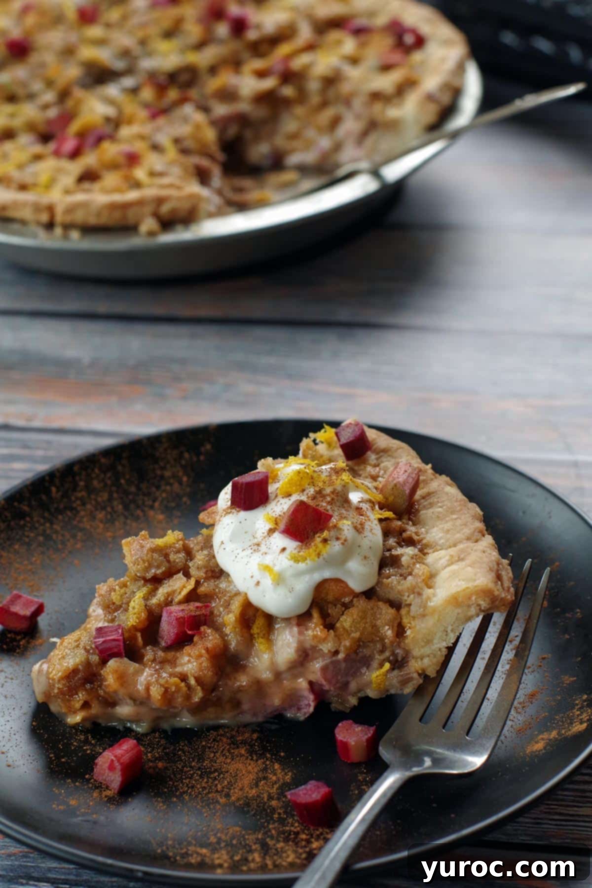 A captivating view of a whole Rhubarb Sour Cream Pie with a slice removed, revealing the delicious interior. A fork and the single slice are elegantly placed on a black plate beside it.