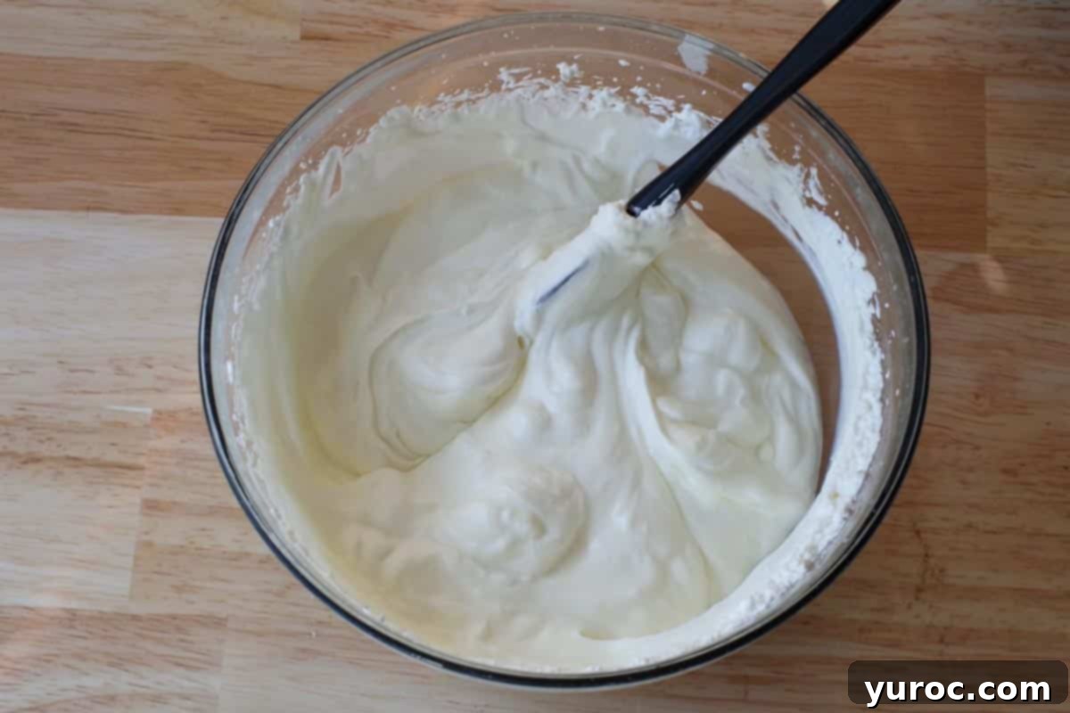 Effortless Butter Tart Dream Ice Cream 10 Sweetened condensed milk mixed into whipped cream in a glass bowl, being gently stirred with a black spatula.