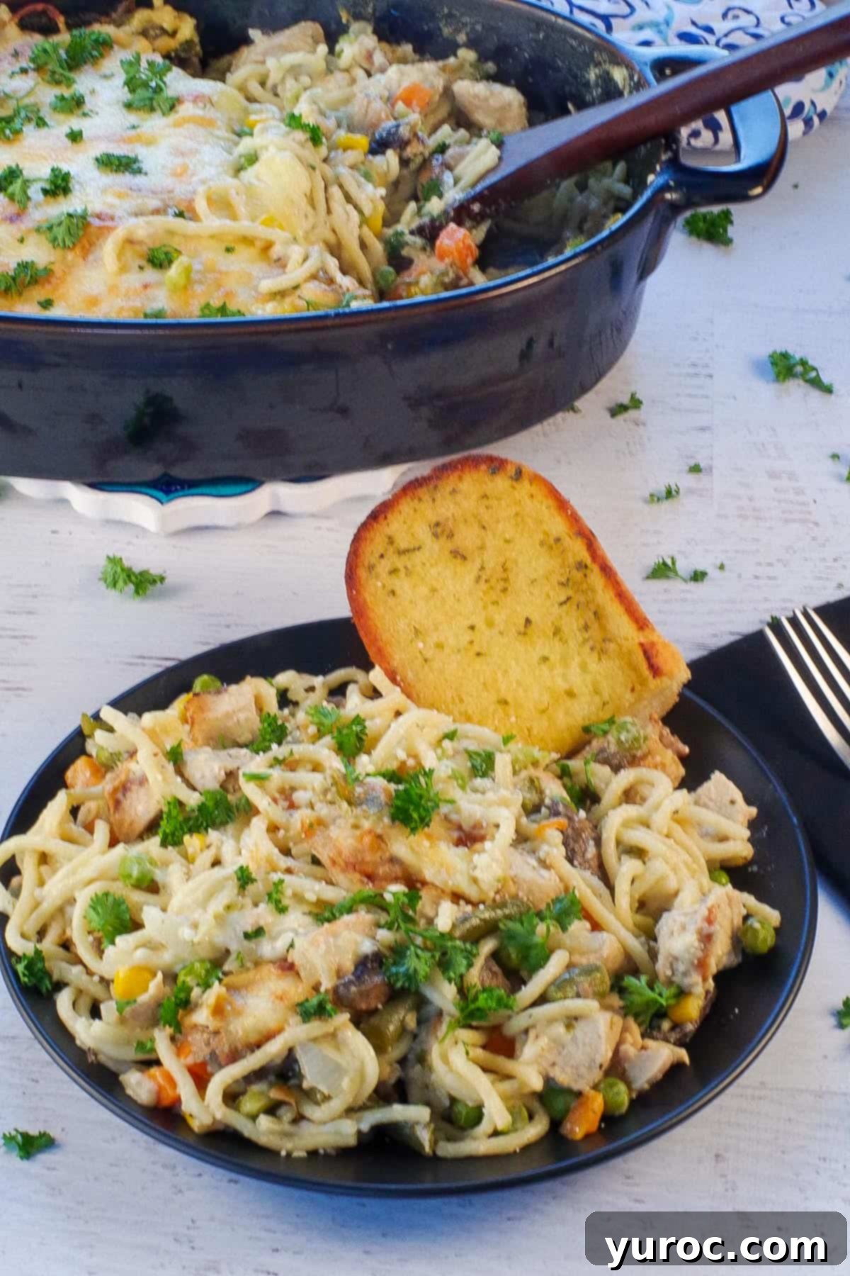 A serving of turkey tetrazzini on a black plate, garnished with parsley, alongside a piece of garlic toast. The full casserole dish is blurred in the background.
