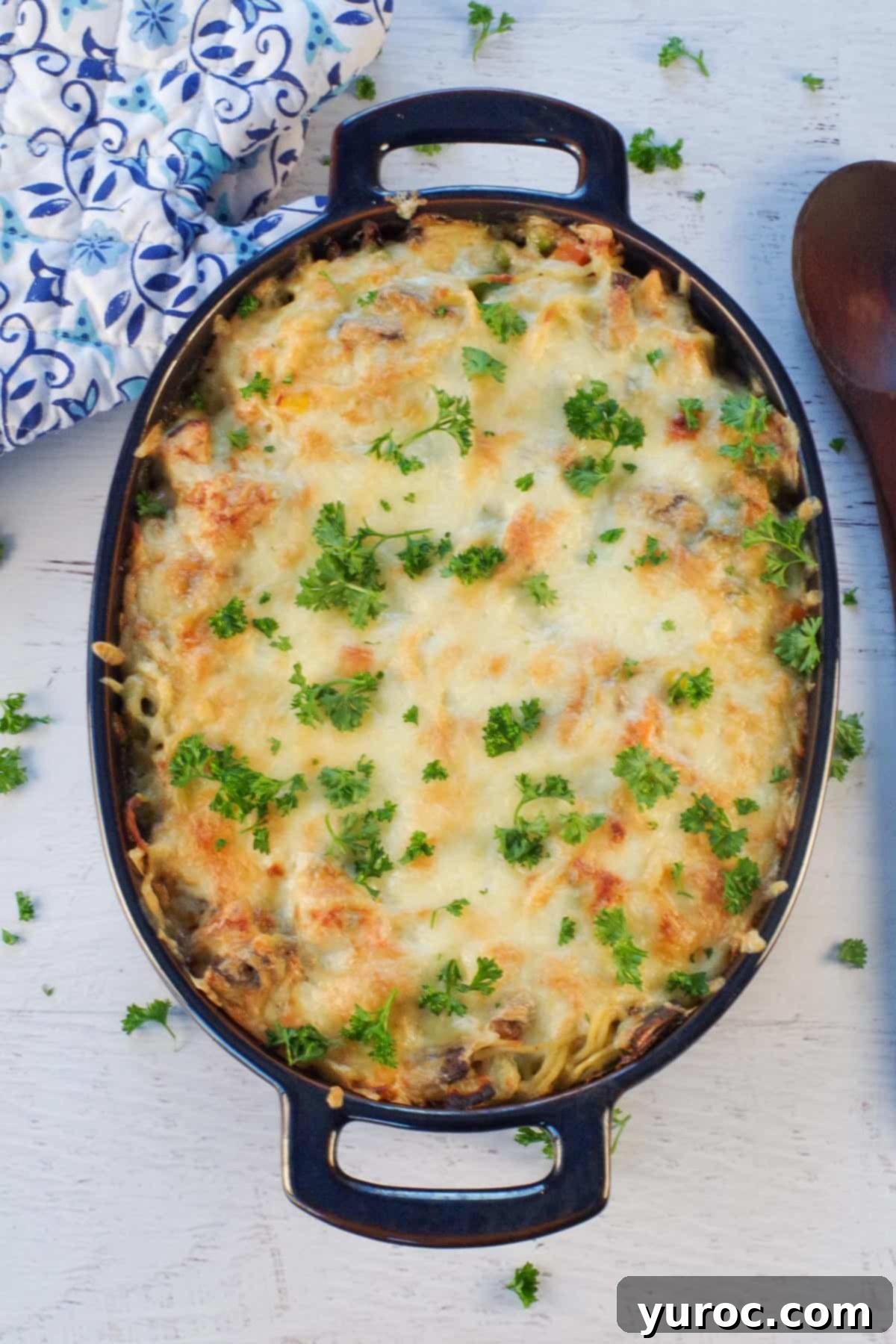 Close-up of baked turkey tetrazzini in a casserole dish, showing the golden cheese and creamy texture, with a wooden spoon and blue oven mitts blurred in the background.