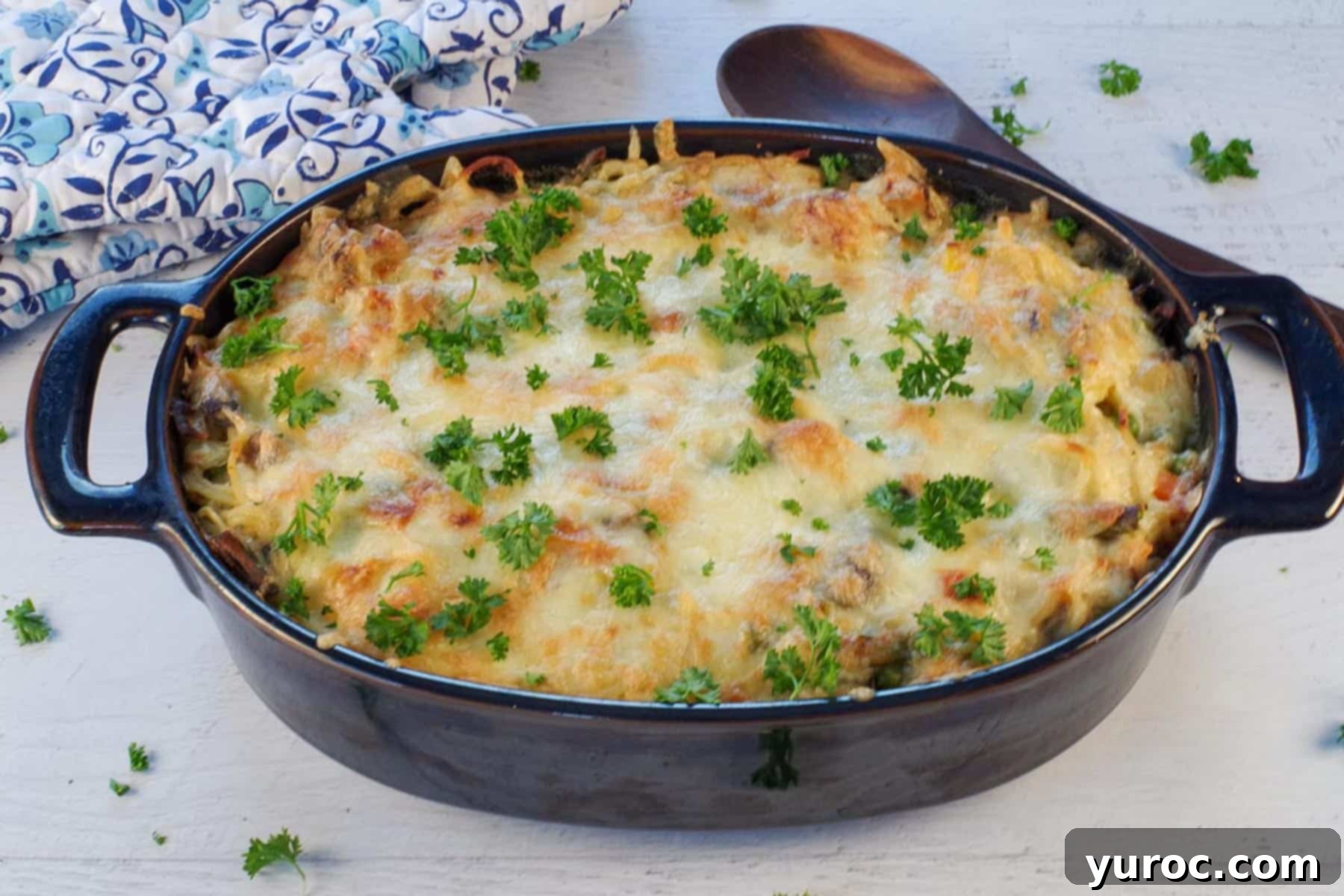 A large casserole dish of freshly baked turkey tetrazzini, steaming and garnished with fresh parsley, with a wooden spoon and blue oven mitts nearby.