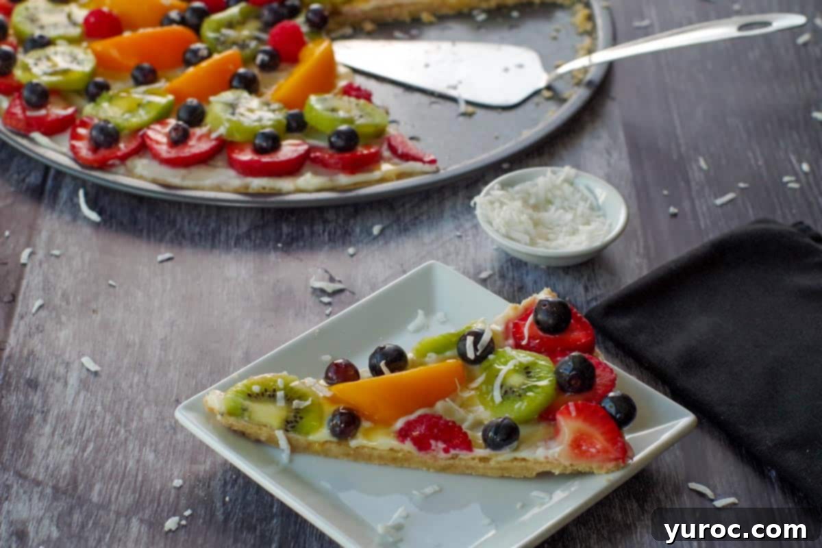 Vibrant Fruit Pizza with Sweet Glaze 2 A perfectly portioned piece of fruit pizza on a white plate, with the remainder of the beautifully decorated fruit pizza in the soft background.