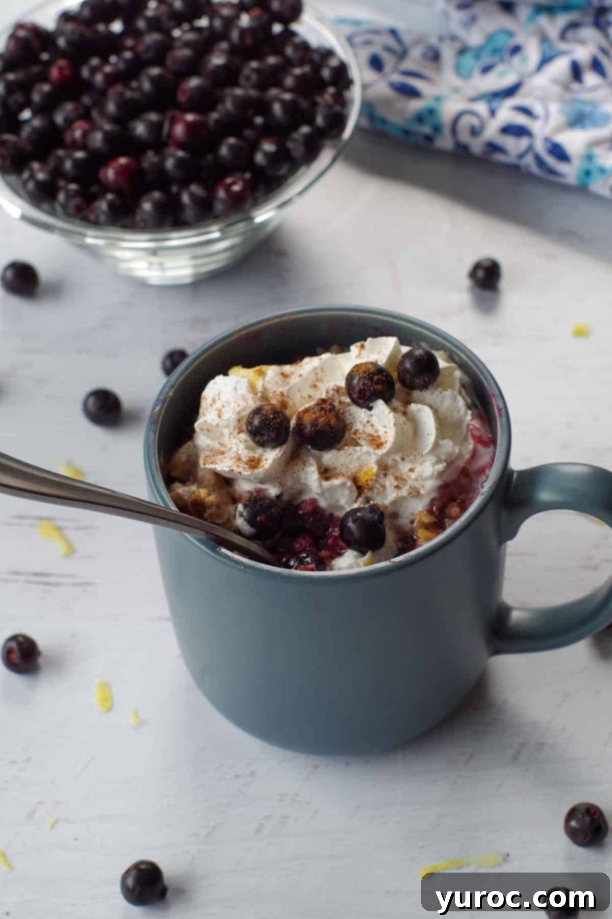 Saskatoon berry crisp recipe in a mug with a spoon in it, berries stewn around and a bowl of Saskatoon berries in the background