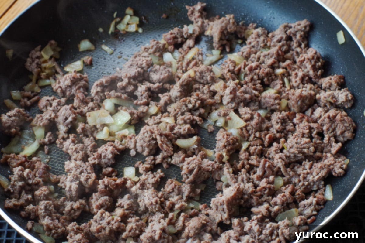 Ground beef cooking in a skillet with chopped onion, ready to be seasoned