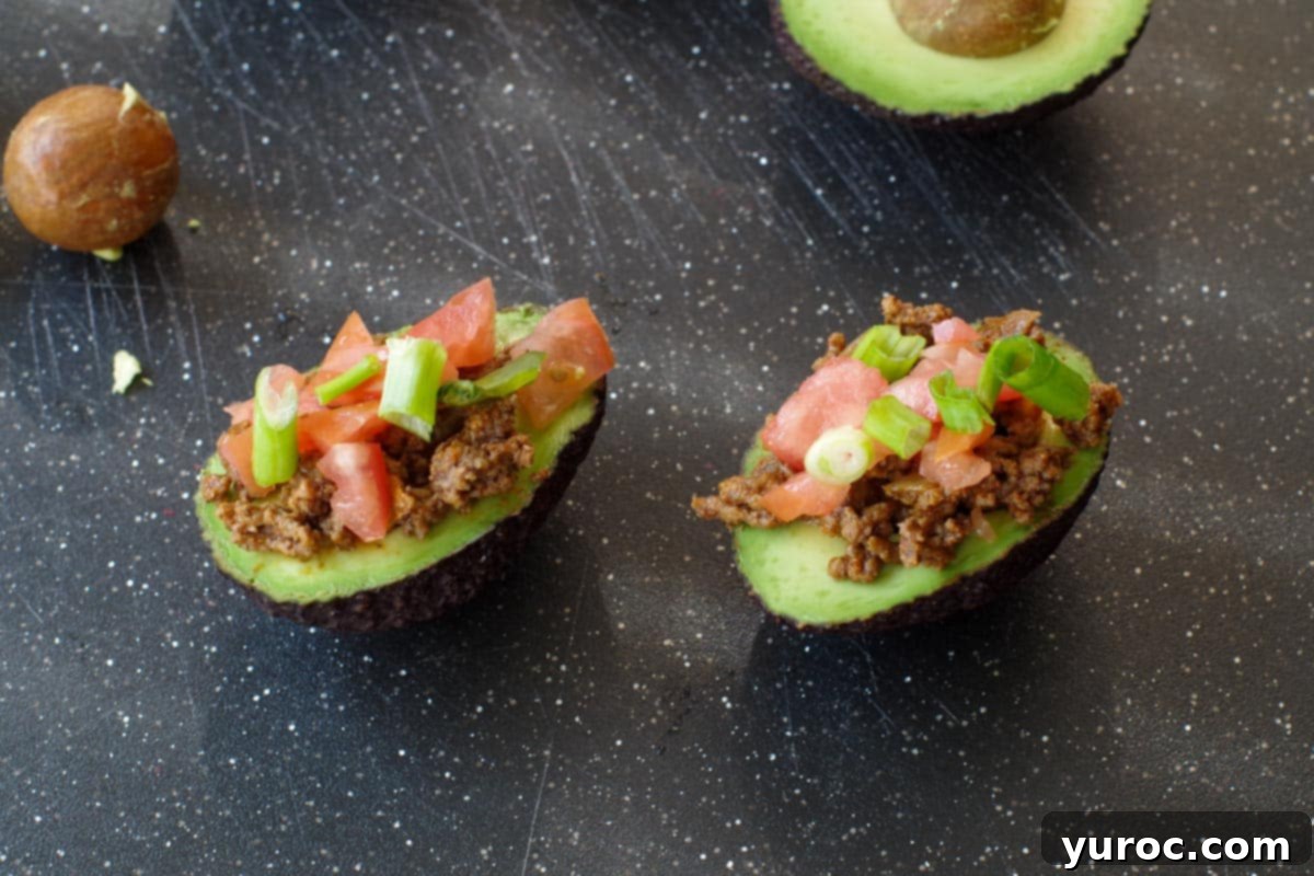 Two prepared avocado tacos on a black cutting board, generously filled with seasoned ground beef, diced tomatoes, and chopped onions