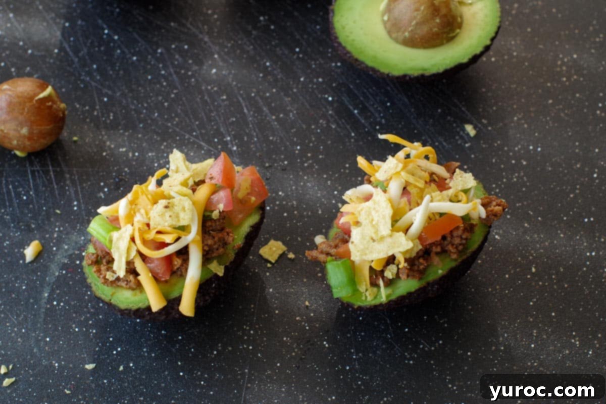 Avocado tacos on a black cutting board, now topped with shredded cheese and crunchy, broken taco shell pieces