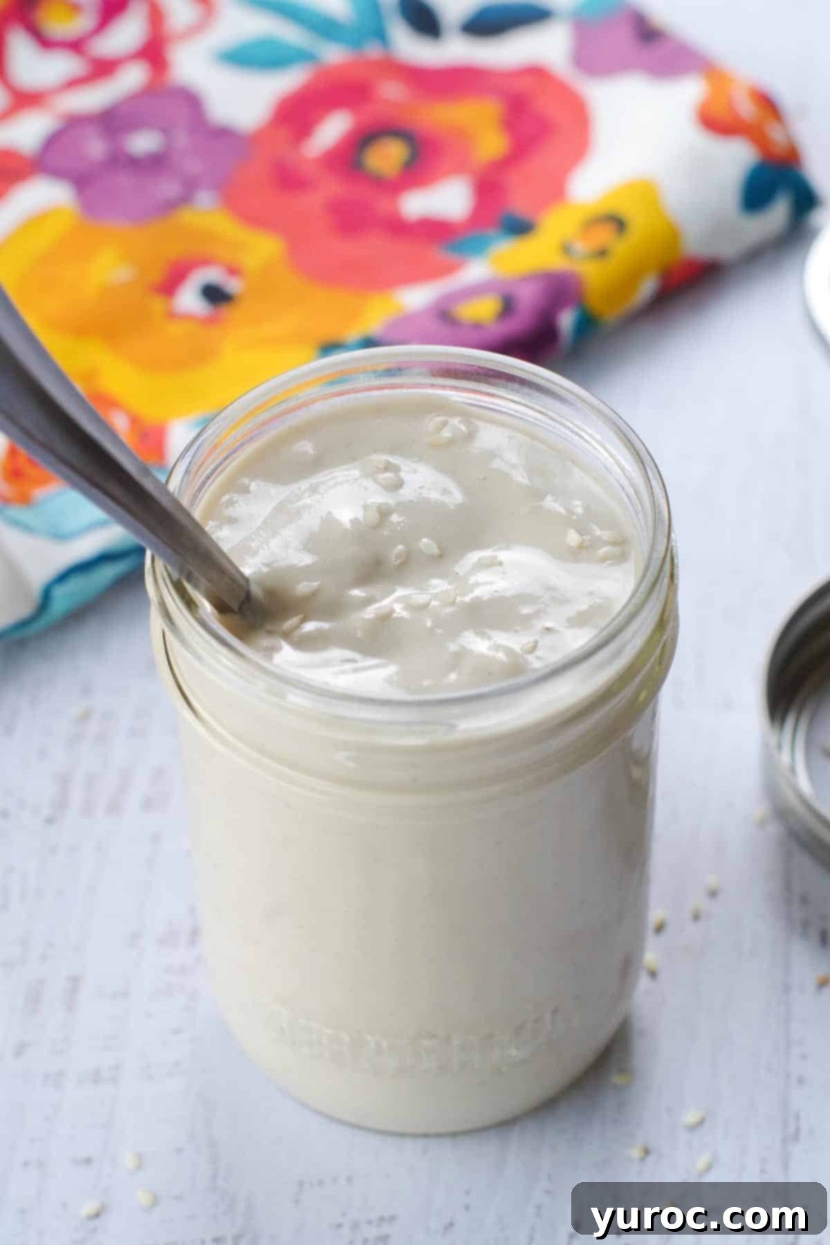 A glass jar filled with creamy, light brown tahini sauce, with a small spoon resting inside it, set against a bright background, showcasing its smooth texture and inviting appeal.