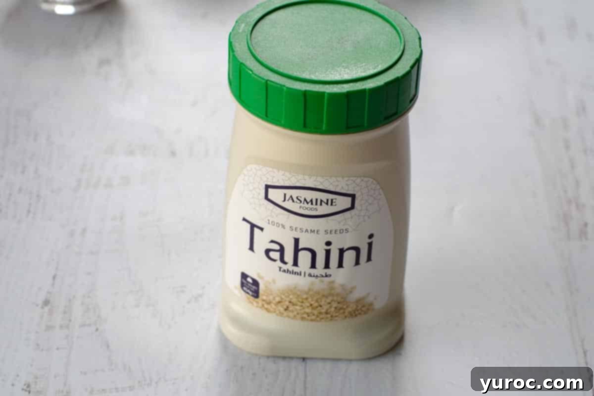 A glass jar of tahini paste (sesame seed paste) sits on a clean white surface, ready to be used as the base for the sauce, emphasizing its pure form before transformation.
