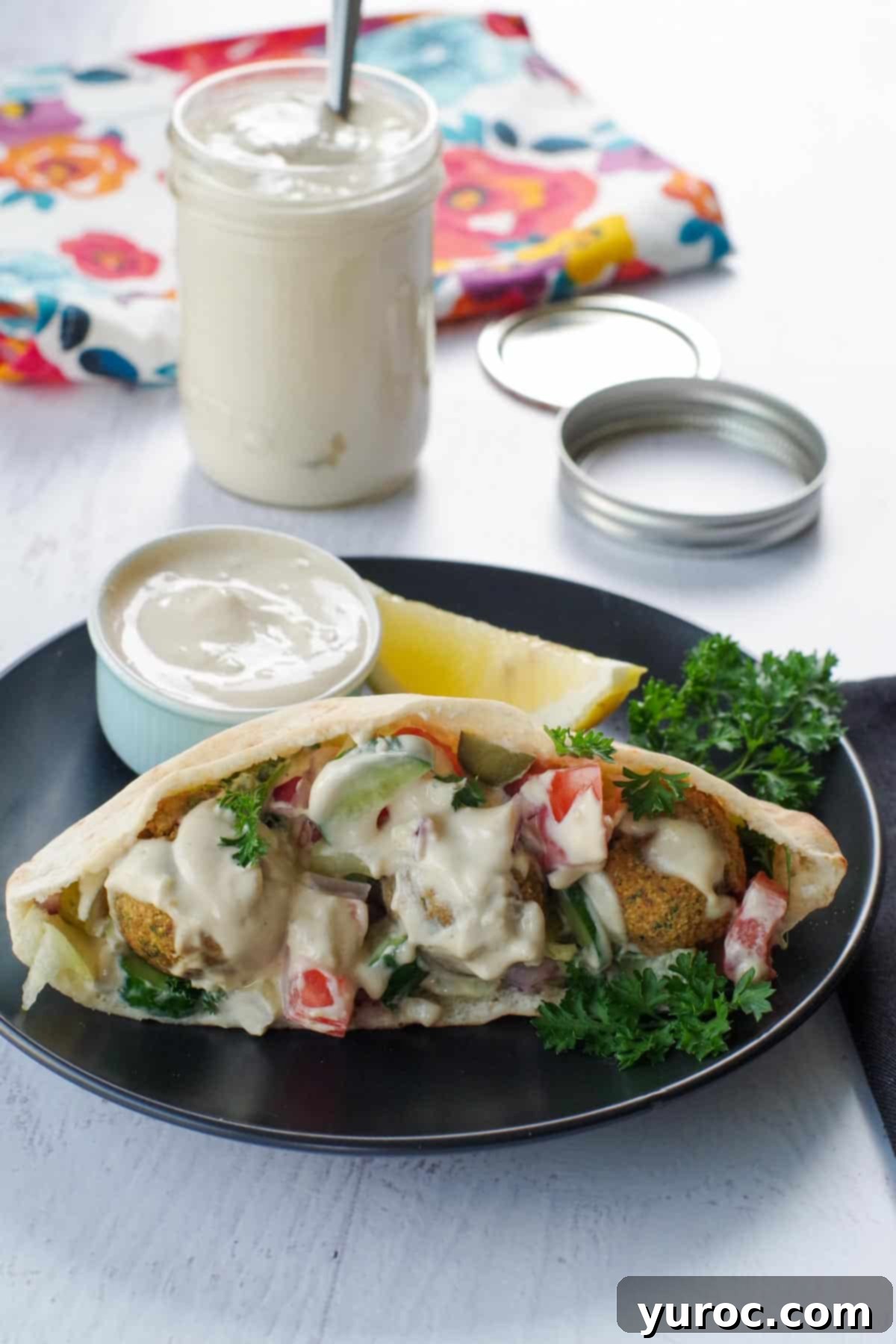 A perfectly cooked falafel rests on a black plate, with a small dish of creamy tahini sauce ready for dipping in the foreground. A jar of tahini paste is blurred in the background, completing the scene, inviting you to enjoy this delicious Middle Eastern meal.