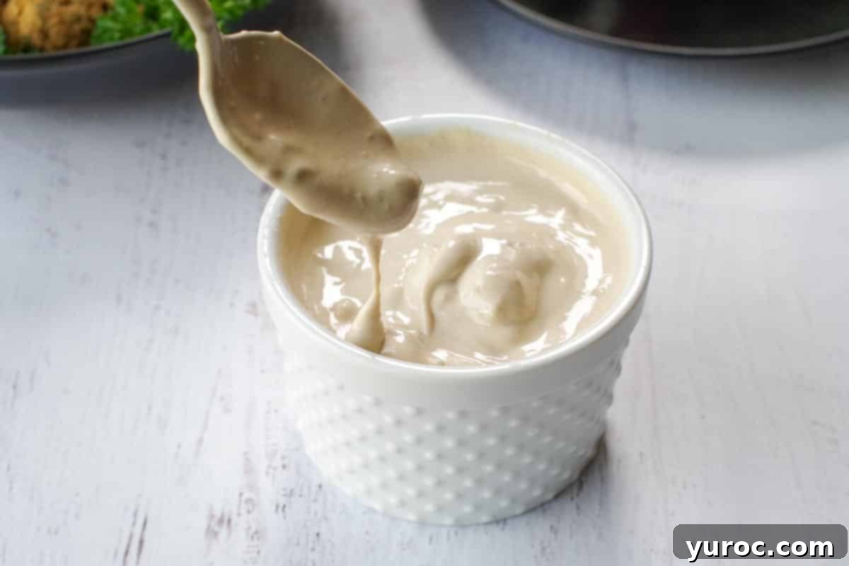 A white ceramic dish filled with creamy tahini sauce, with a spoon gently scooping some out, highlighting the smooth texture and ready-to-serve appeal, set on a dark surface.