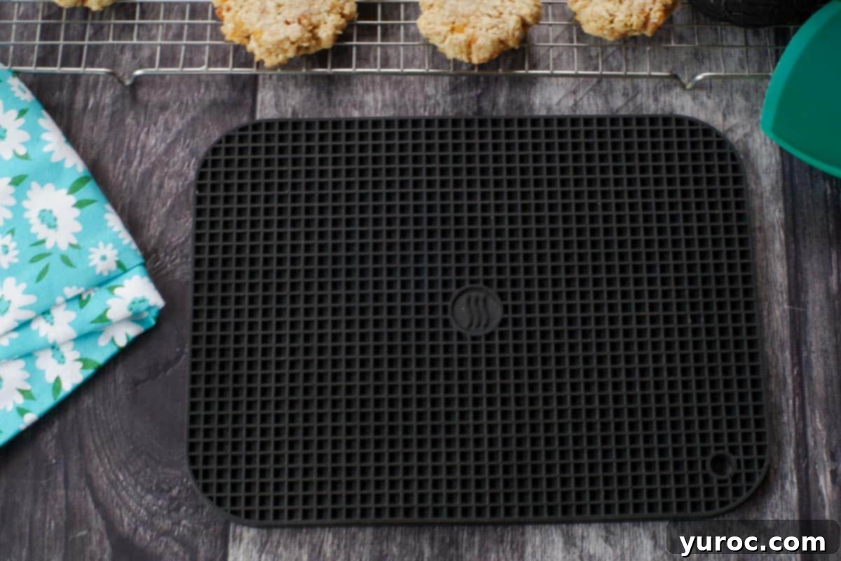 Large black silicone trivet on a faux wood surface with freshly baked cookies in the background, showcasing its utility