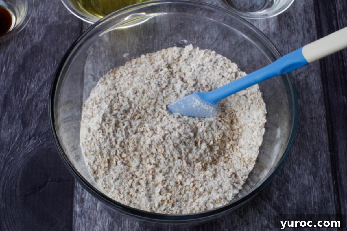 Dry ingredients, including flour, oats, and shredded coconut, combined in a large glass bowl with a blue spatula nearby