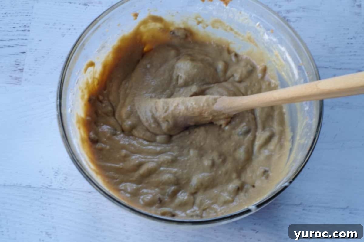 Milk and raisins added to cupcake batter in a large glass bowl, with a wooden spoon