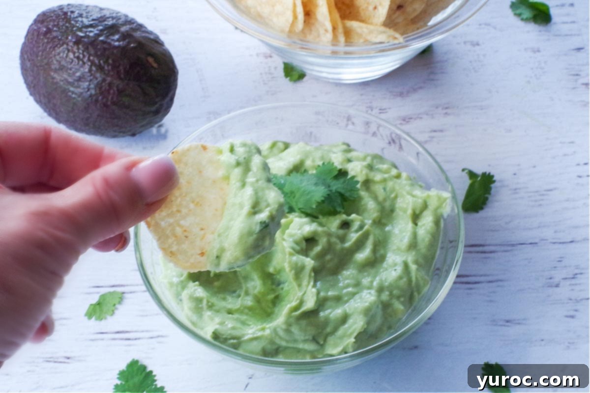 avocado cream sauce in a glass bowl with a tortilla chip being dipped in it, and a bowl of tortilla chips in the background