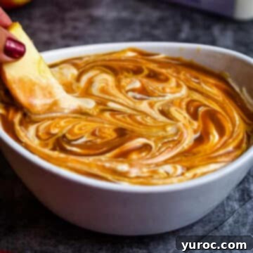 A luscious caramel apple cheesecake dip served in a pristine white bowl, with a slice of apple being dipped into its creamy goodness.