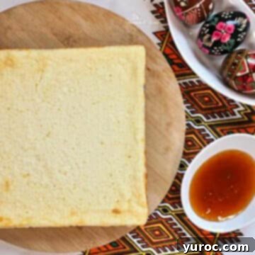 A square Ukrainian cheesecake, known as Easter Syrnyk, elegantly presented on a round wooden serving board, with a side of sauce and a bowl of traditional Ukrainian Easter eggs in the background.