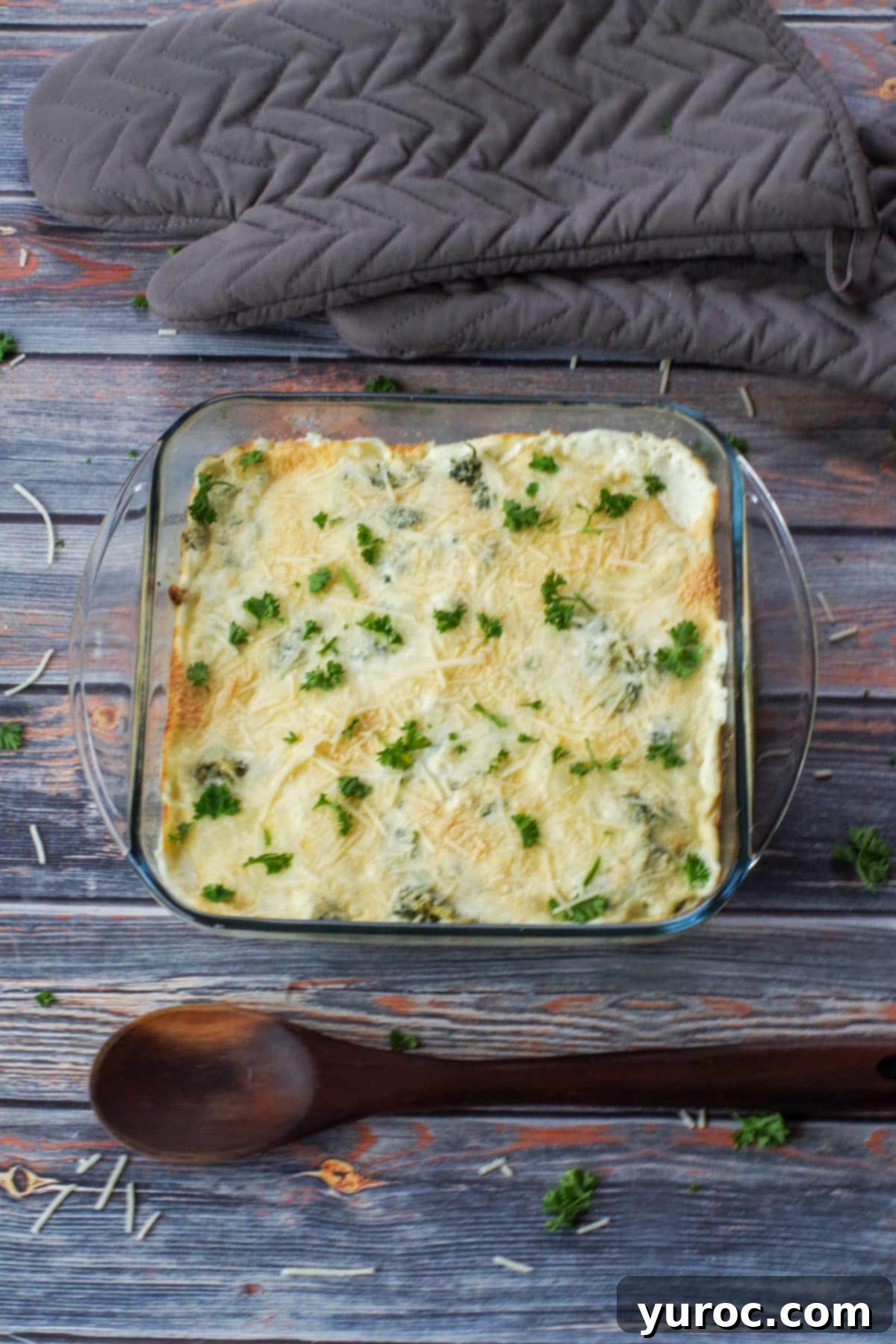 spinach artichoke casserole with wooden spoon below it and grey oven mitts above casserole
