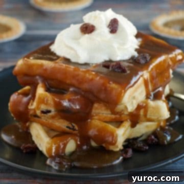 A stack of four golden butter tart waffles, generously drizzled with butter tart sauce, presented on a sleek black plate, with more butter tarts visible in the background