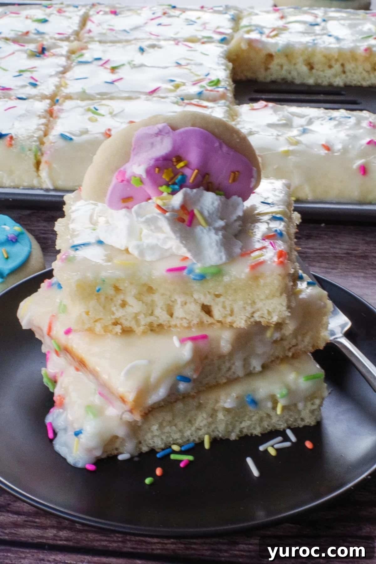 a stack of 3 sugar cookie sheet pan pancakes on a black plate, garnished with half a sugar cookie and whipped cream, with a pan of more pancakes in the background