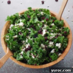 Kale Cranberry and Feta salad in a wooden bowl with wooden utensils and cranberries and sunflower seeds strewn around