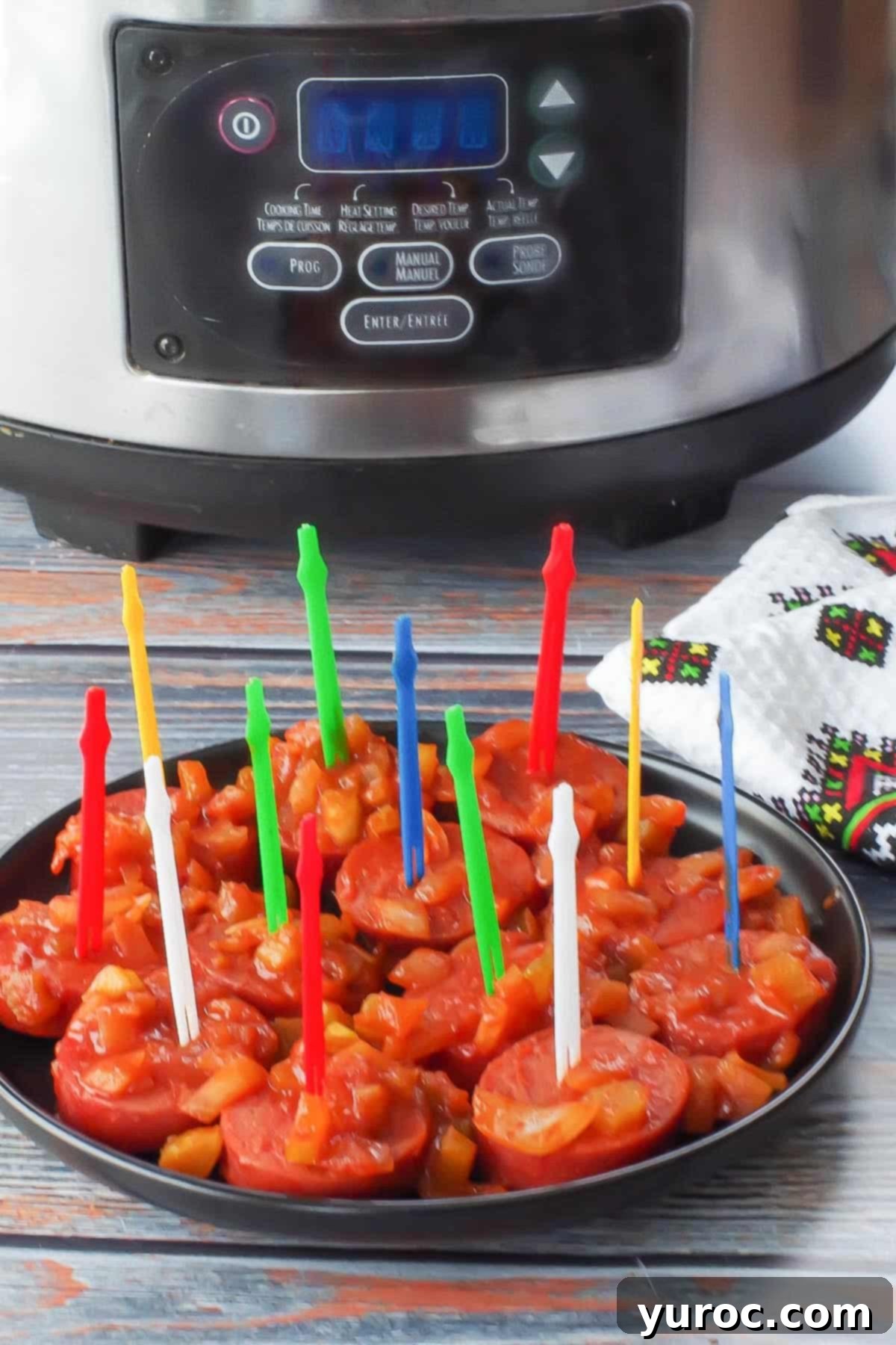Slow cooker kielbasa appetizer with colorful plastic skewers, on a black round plate with a slow cooker in the background, ready to serve.