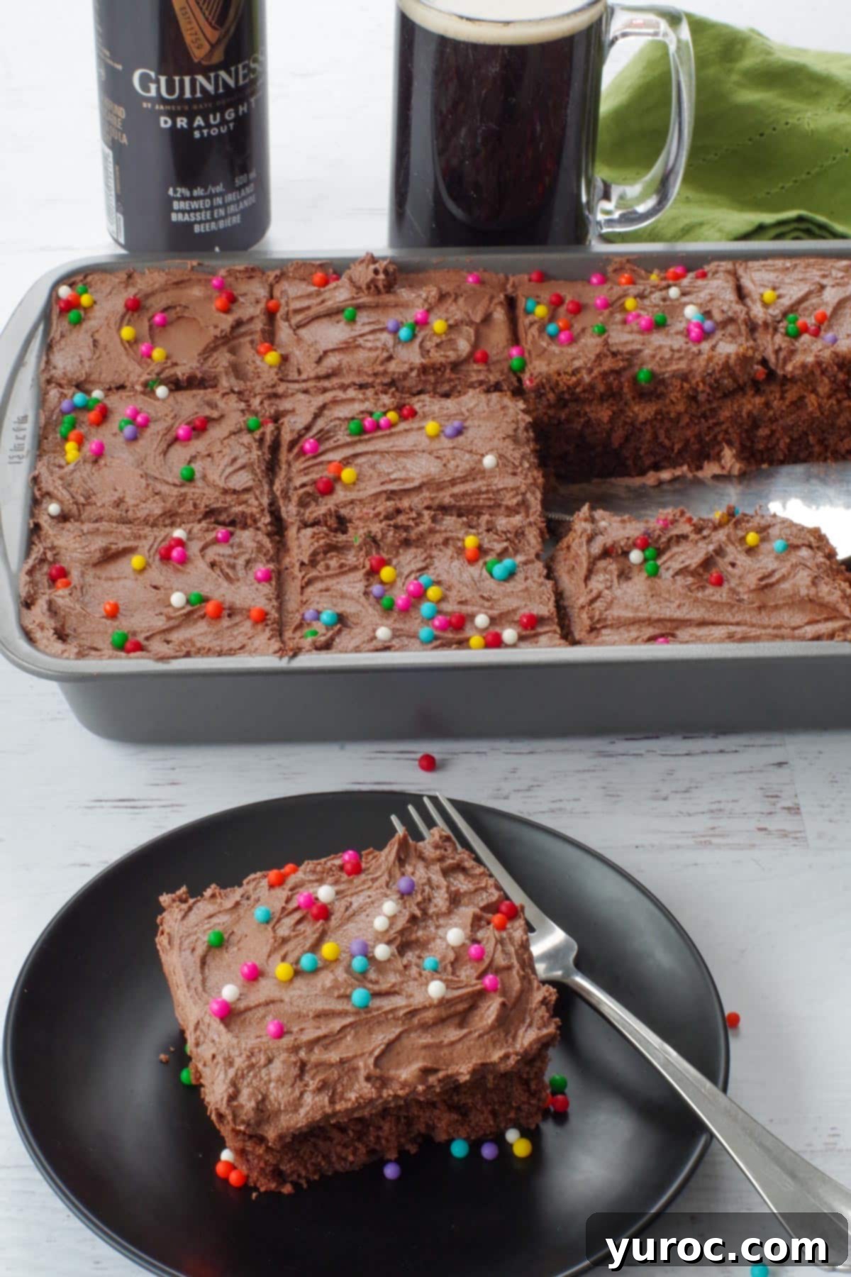 A slice of rich chocolate Guinness cake with creamy Irish cream frosting on a black plate, with the rest of the cake in a pan in the background.