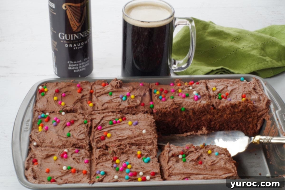 A 9x13 inch pan of chocolate Guinness cake, with a glass of Guinness beer and a can of Guinness in the background, set on a rustic wooden surface.