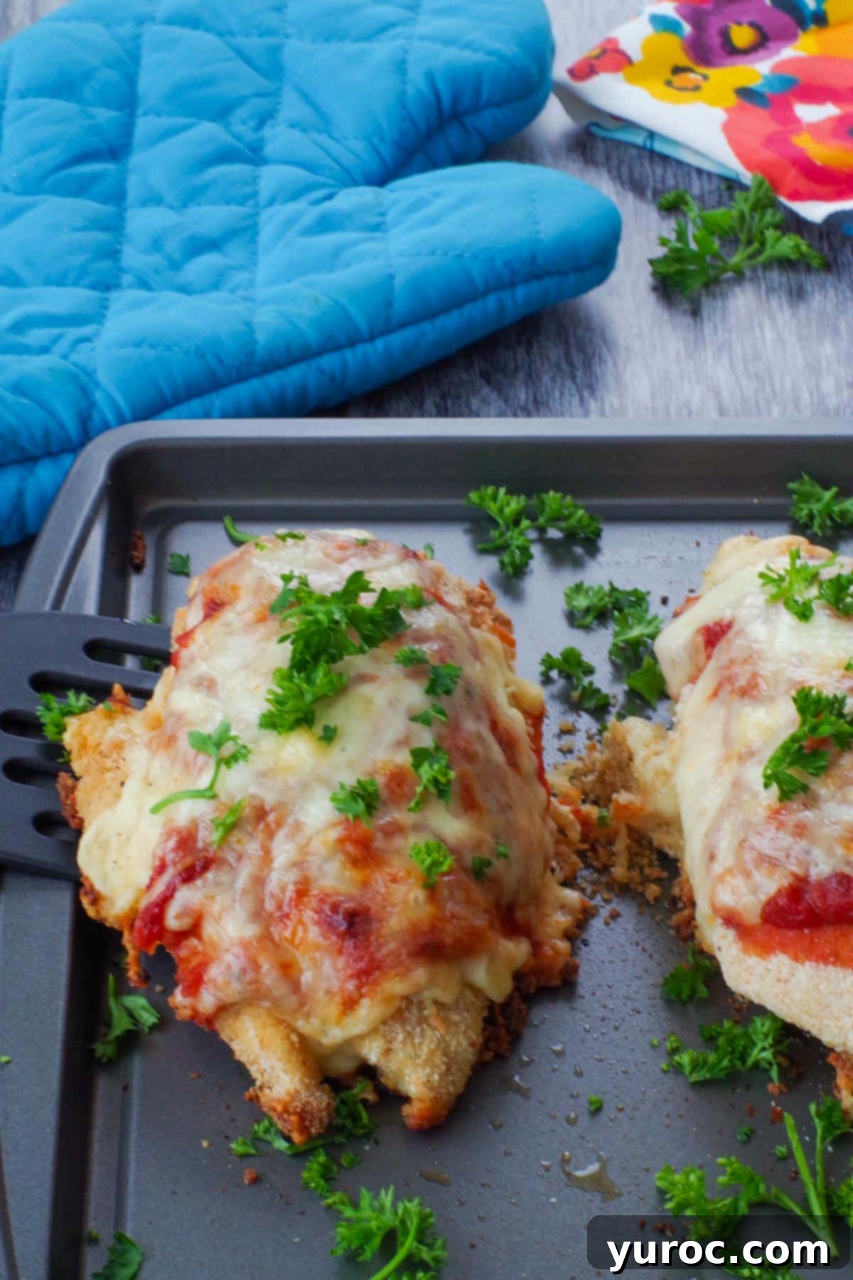 Two golden-brown chicken parmesan breasts on a sheet pan, ready to serve, with a blue oven mitt in the background.