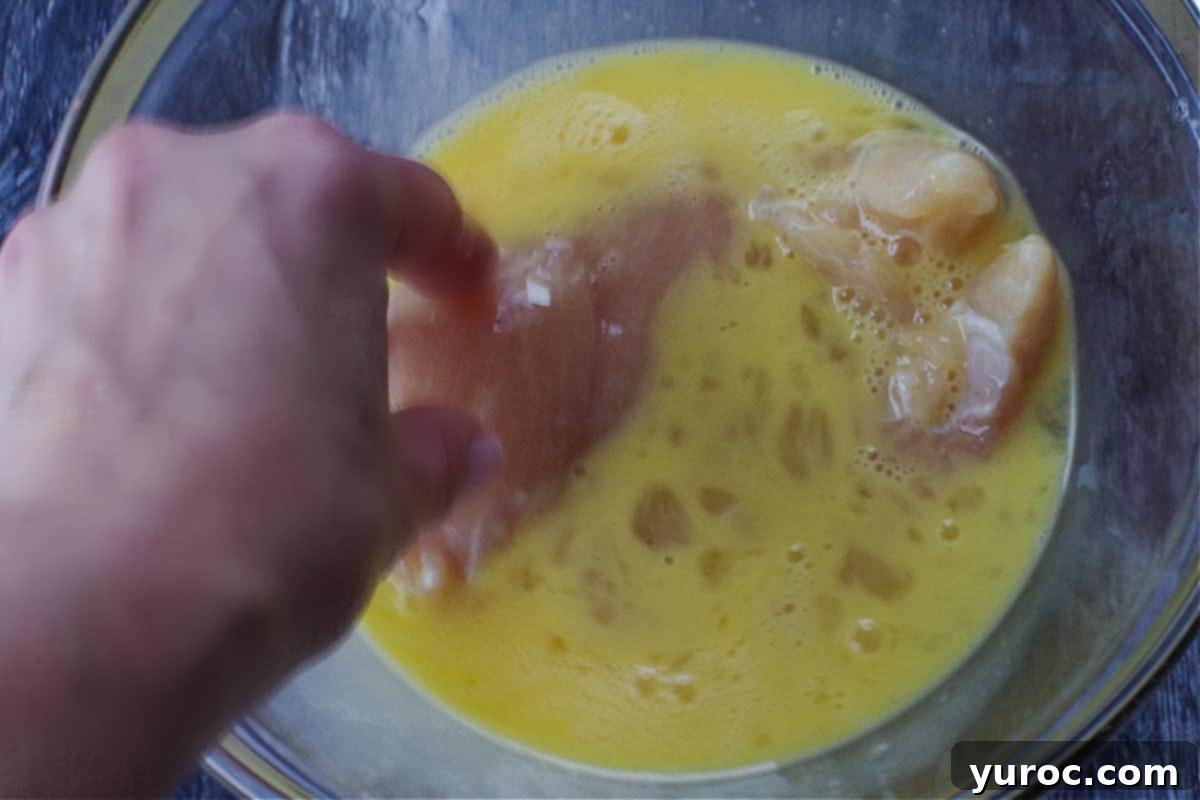A chicken breast being dipped into the egg mixture in a glass bowl.