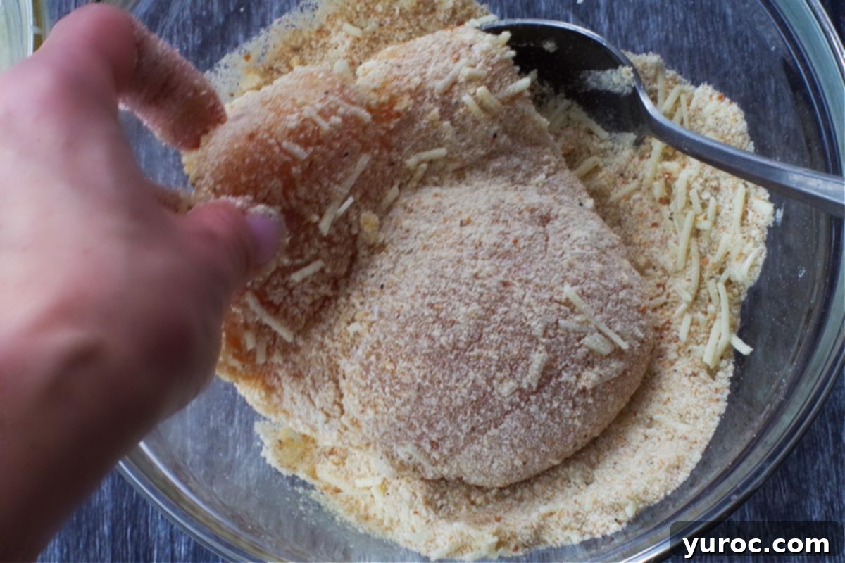 An egg-coated chicken breast being pressed into the bread crumb mixture to create an even coating.