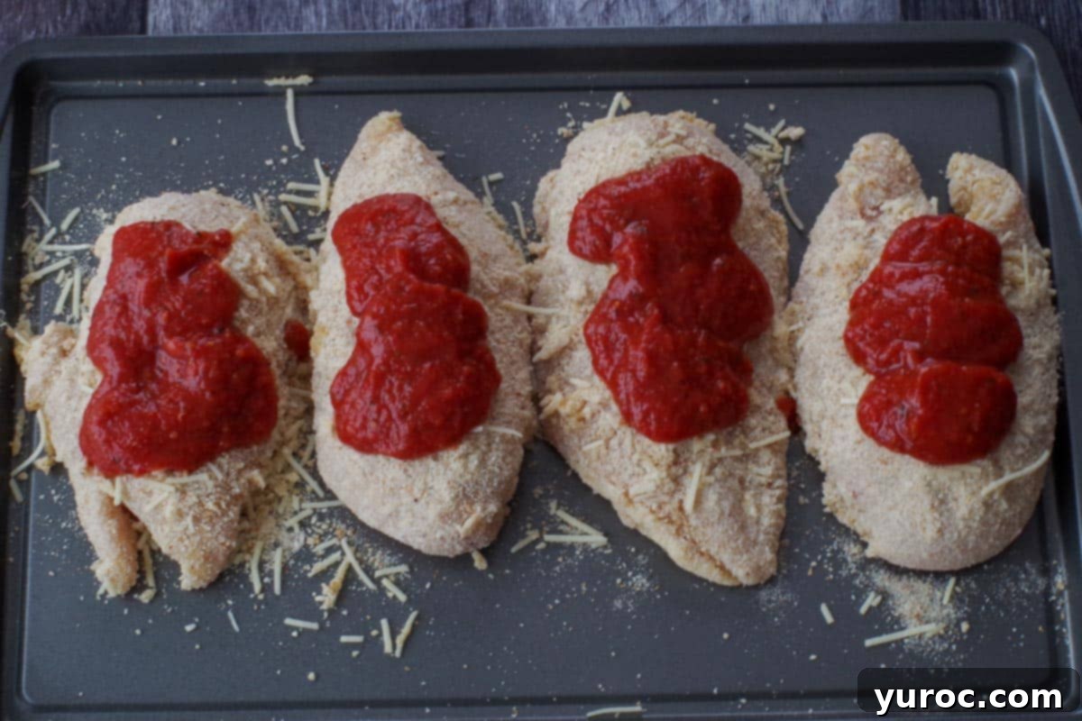 Pasta sauce being spooned over the top of the breaded chicken breasts on the baking sheet.