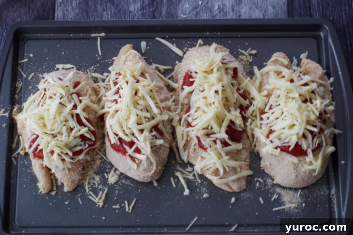 Shredded mozzarella cheese generously sprinkled over the sauced chicken breasts on the baking sheet.
