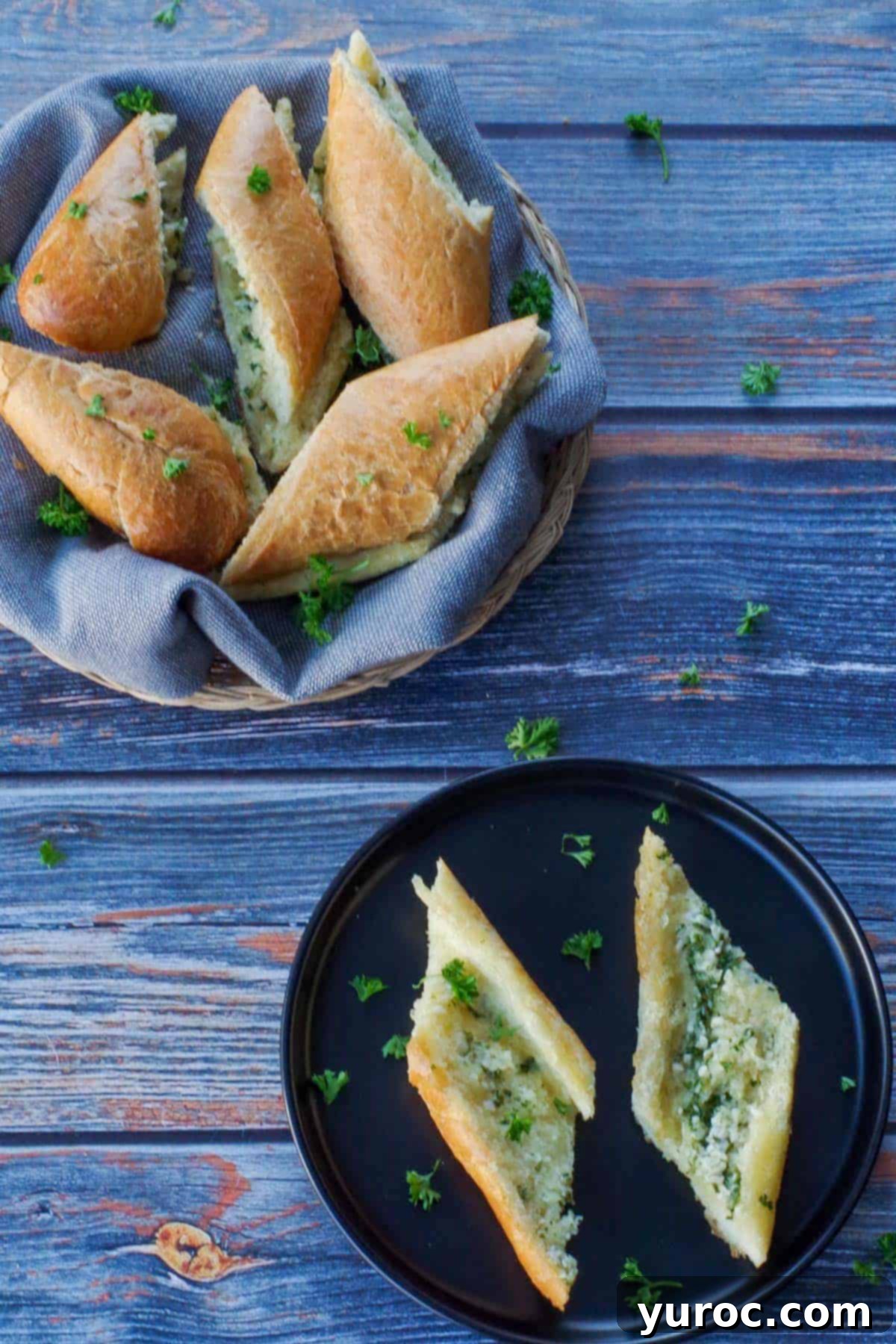 Two golden-brown slices of garlic bread rest on a sleek black plate, with a basket of additional sliced baguette garlic bread in the background, showing off its crispy crust and soft interior.