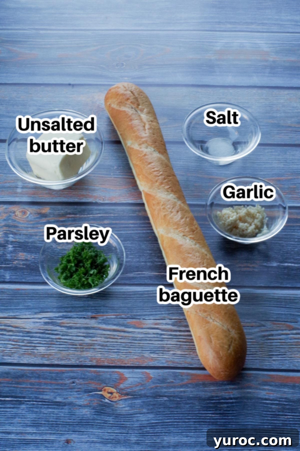 Fresh ingredients for garlic bread baguette, including a whole baguette, softened butter in a glass bowl, fresh parsley, and garlic cloves, all arranged on a blue faux wooden surface.
