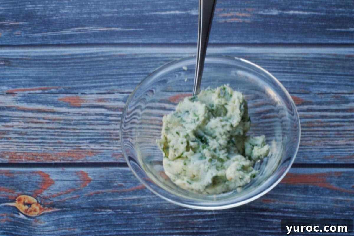 Softened butter, minced fresh parsley, minced fresh garlic, and salt are perfectly mixed together in a small clear glass bowl, with a spoon resting in the mixture.
