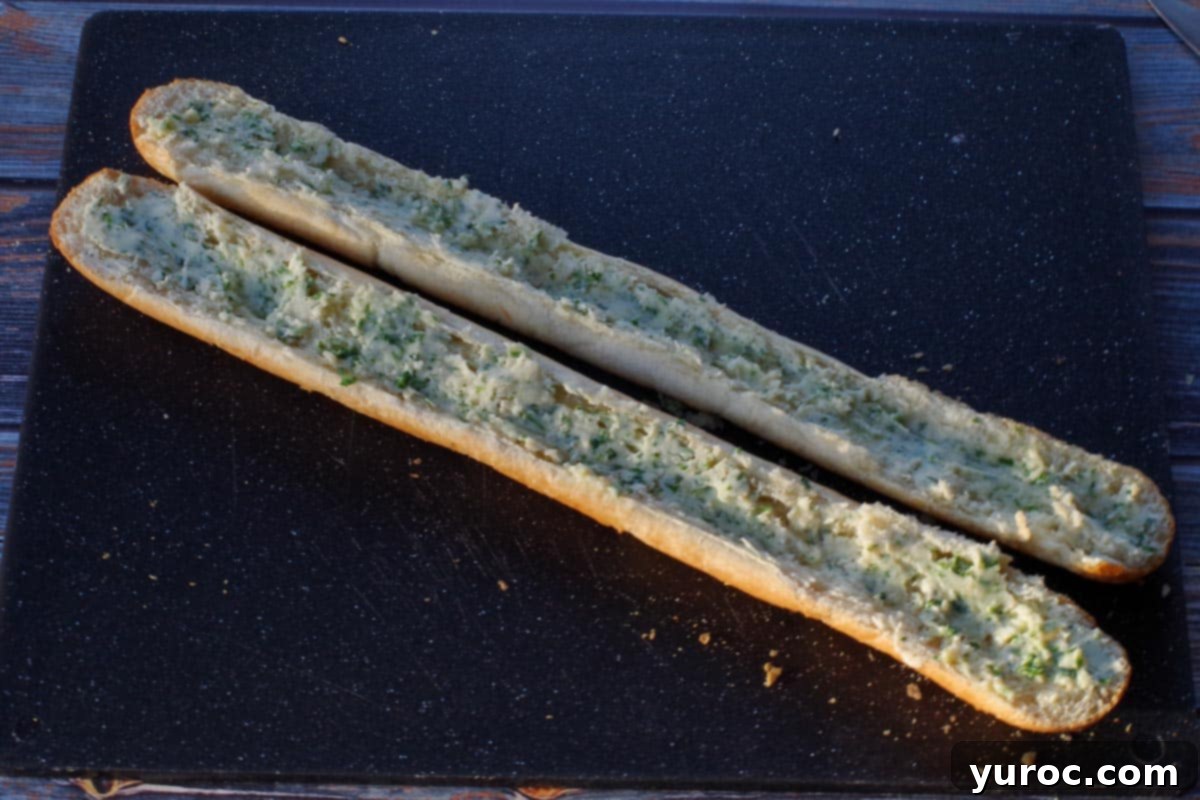 A baguette is cut in half horizontally, revealing its soft interior. The prepared garlic butter mixture is being evenly spread onto the cut sides of both baguette halves, resting on a black cutting board.