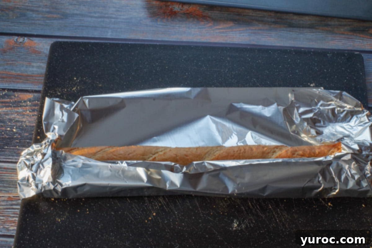 The aluminum foil is being folded over the short side of the buttered baguette first, demonstrating the proper wrapping technique on a black cutting board.