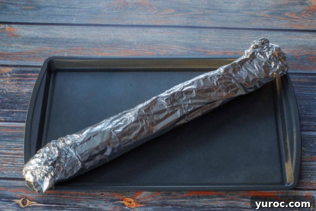 The baguette is tightly wrapped in aluminum foil, creating a sealed packet, and placed on a baking sheet, ready for the oven.