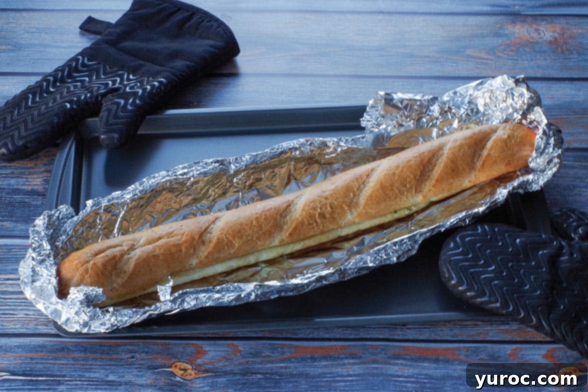 A perfectly baked, foil-wrapped baguette is shown, having just been removed from the oven, indicating it's ready for careful unwrapping.