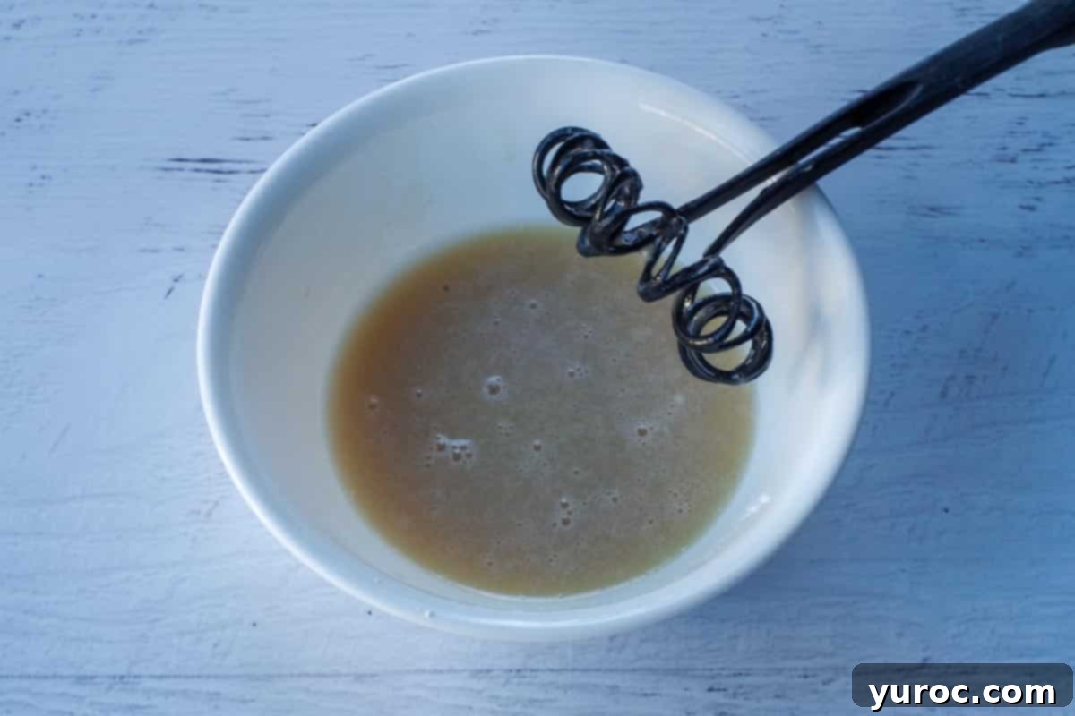 Ingredients for the glaze, powdered sugar, vanilla coffee, and milk, whisked together in a white bowl with a black whisk, creating a smooth, luscious mixture.