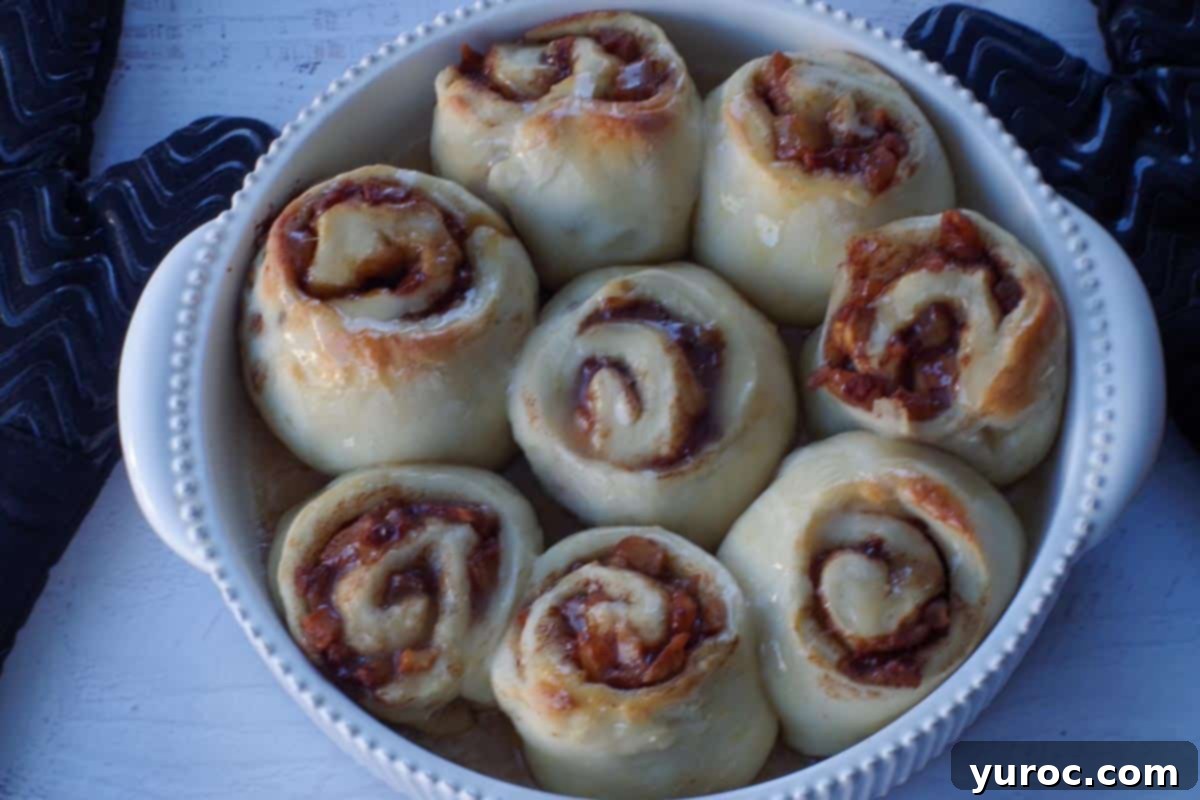 The delicate glaze is being poured generously over the warm, freshly baked cinnamon rolls, coating them in a sweet sheen.