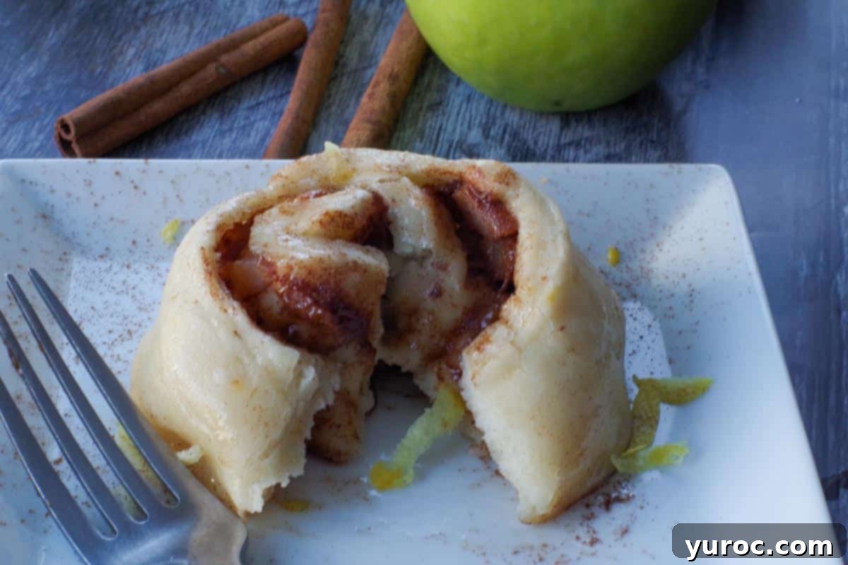 A single apple pie cinnamon roll, freshly cut open, sits invitingly on a white plate, garnished with a sprinkle of lemon zest and cinnamon sticks in the background, alongside a vibrant green apple.