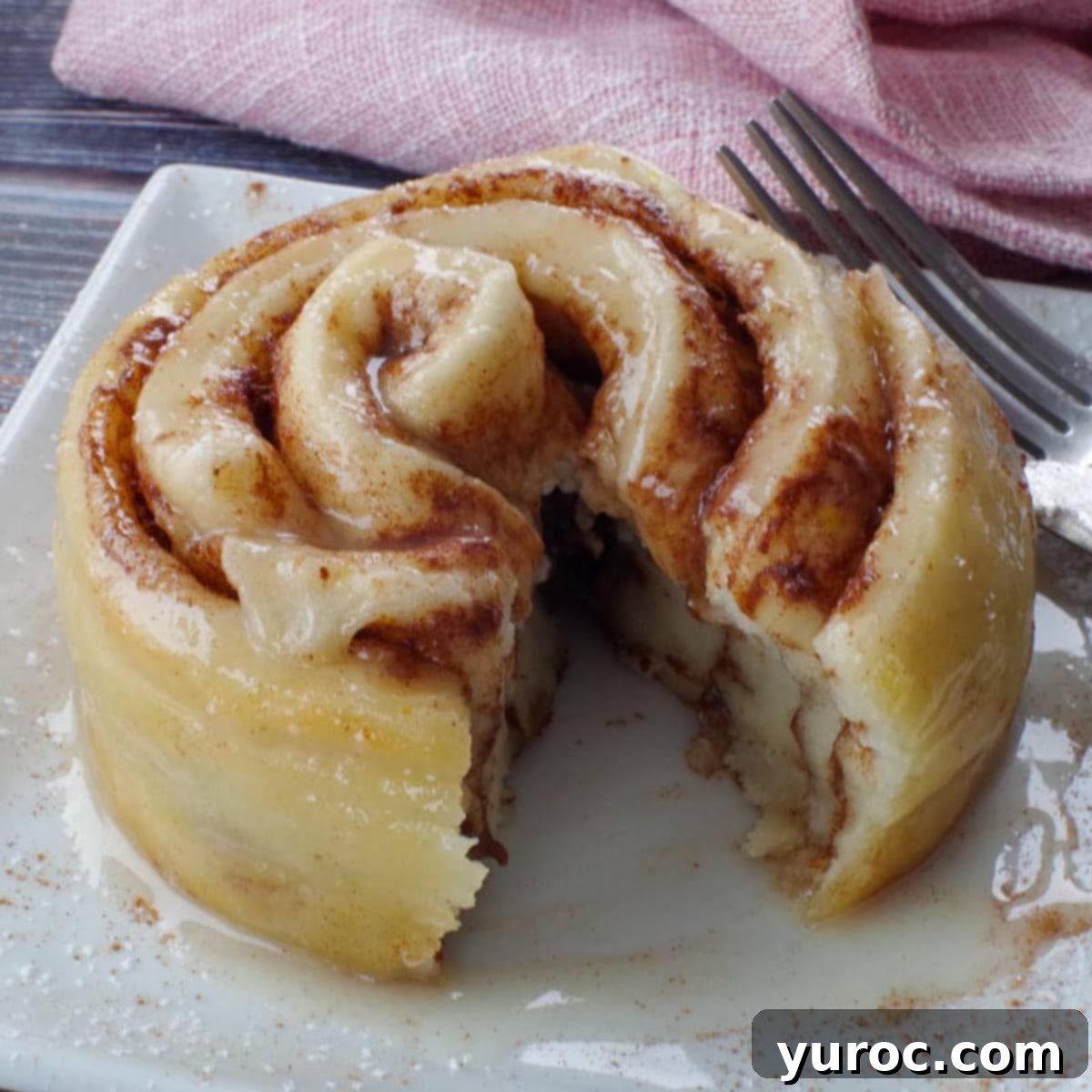 A healthy cinnamon roll in a mug, cut open to reveal its layers, artfully placed on a white plate with a soft pink napkin in the background, making it an inviting single serving.