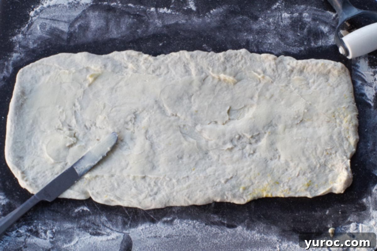 Two-ingredient dough rolled into a perfect rectangle on a large cutting board, with softened light butter spread evenly across its surface.