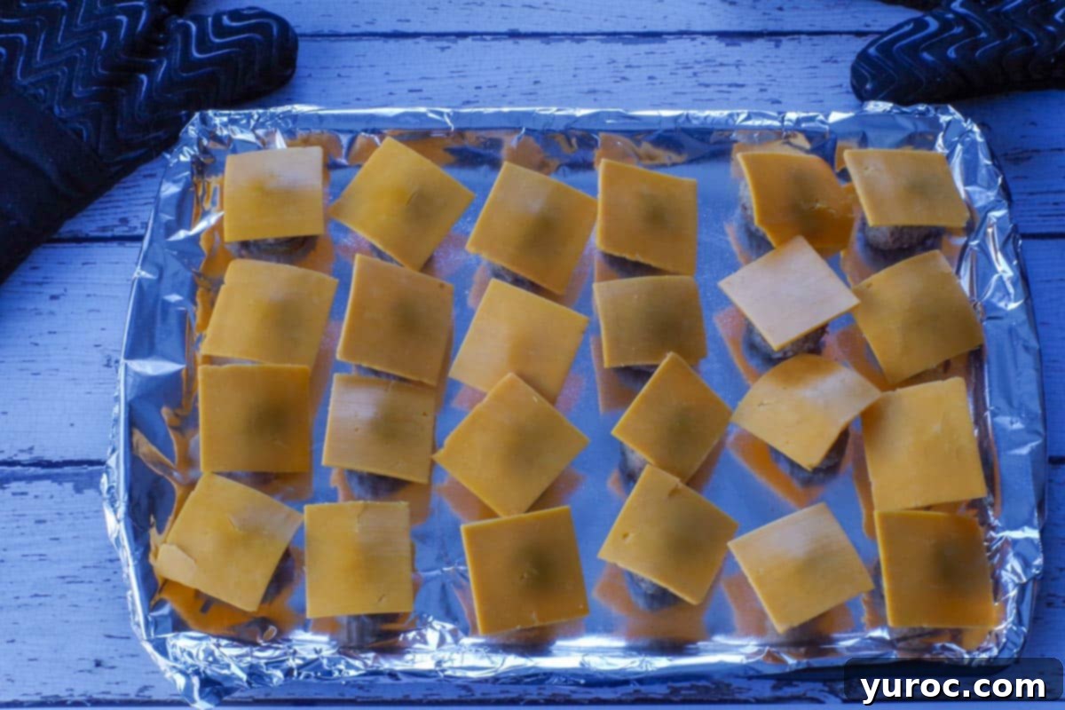 Each cooked meatball on the baking sheet now has a quarter-slice of cheddar cheese neatly placed on top, ready for melting.