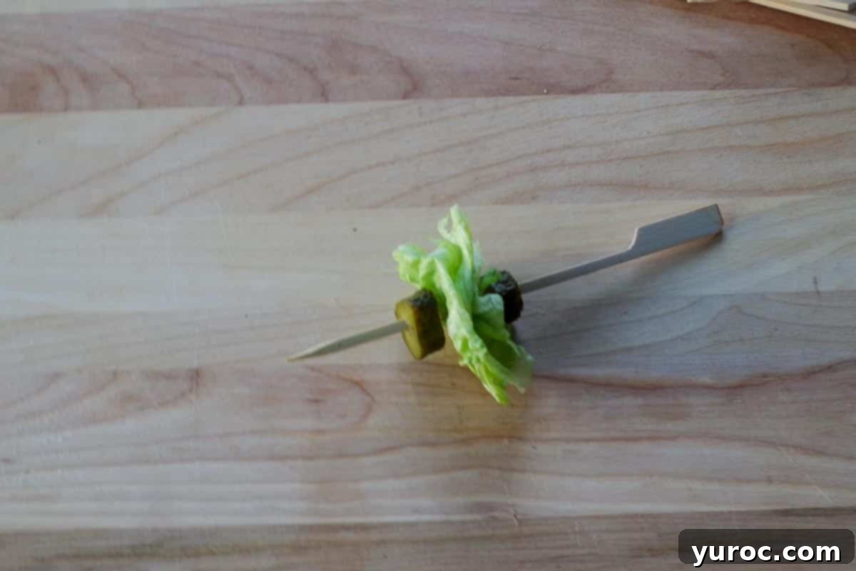 A bamboo skewer with a sequence of pickle slice, shredded lettuce, and another pickle slice, illustrating the base assembly for the Big Mac Bites.