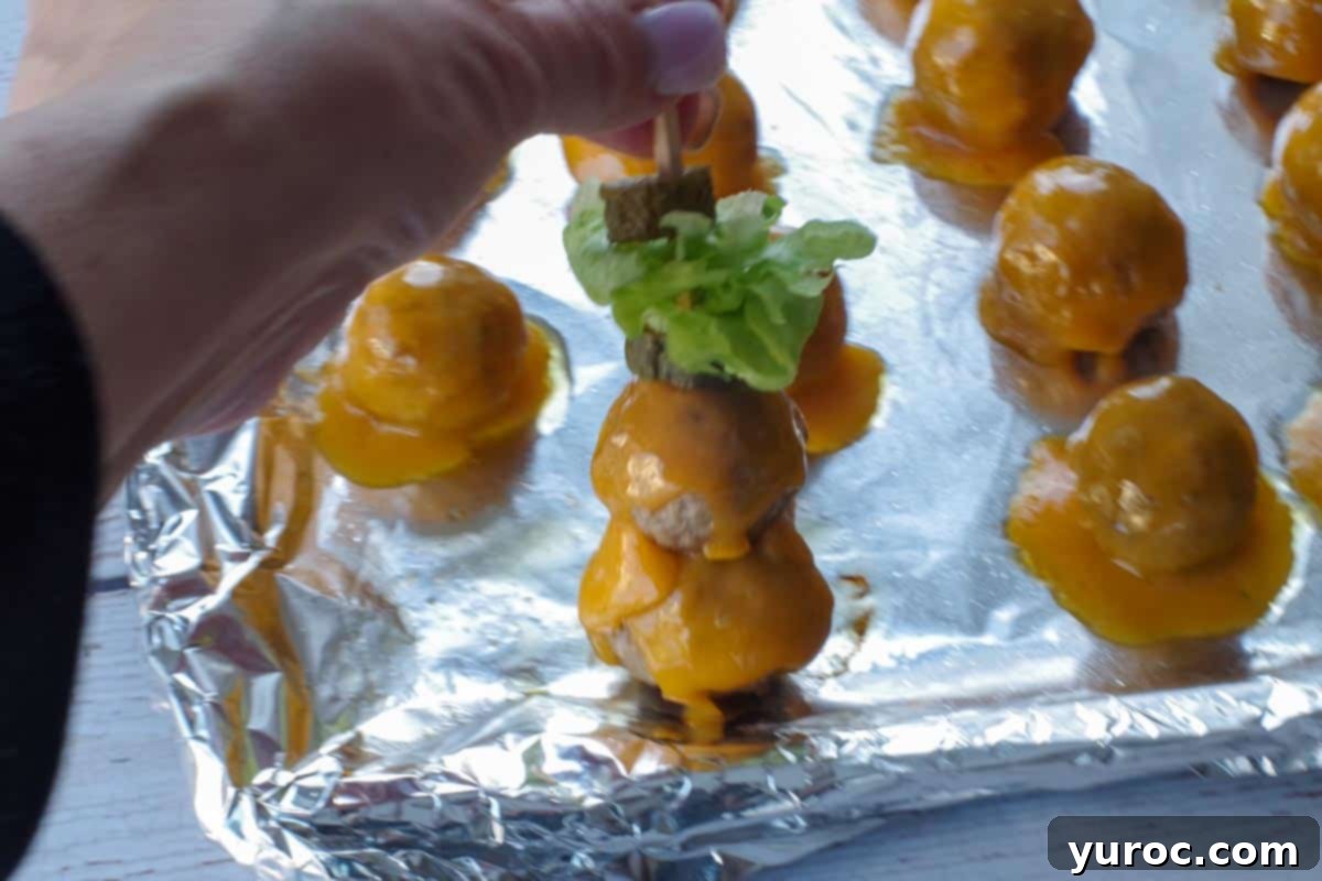 Two cheesy meatballs are being carefully added to the already prepared skewer, positioned above the pickle and lettuce layers.