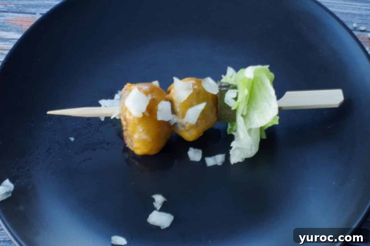 A completed Big Mac Bite skewer, topped with chopped onion, resting on a black plate, ready for sauce.