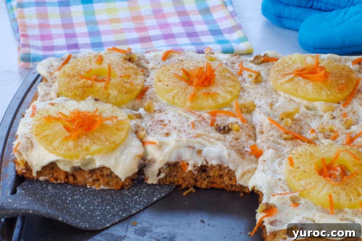 A perfectly portioned carrot cake pancake being lifted off a sheet pan, showcasing its fluffy texture and golden-brown edges. In the background, a colorful blue, yellow, and pink checkered tea towel and blue oven mitts add a touch of warmth to the scene.
