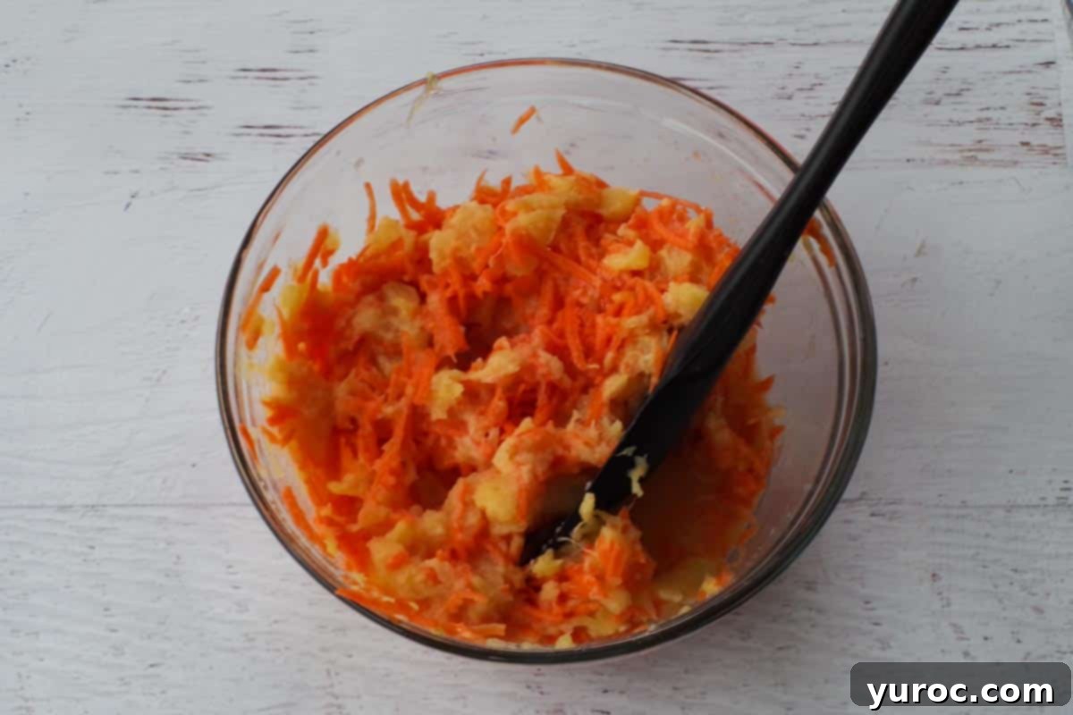 A colorful mixture of fresh carrots, crushed pineapple, unsweetened applesauce, and vanilla extract are whisked together in a medium glass bowl, forming the wet ingredient base for the carrot cake pancakes.